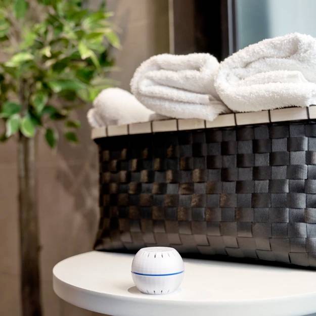 Woven basket filled with white towels on a table, with a round diffuser beside it. Blurred plants in the background.