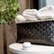Woven basket filled with white towels on a table, with a round diffuser beside it. Blurred plants in the background.