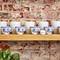 A wooden rack holding ten teacups in an Asian style, positioned in front of a brick wall. A plant is situated in the foreground.