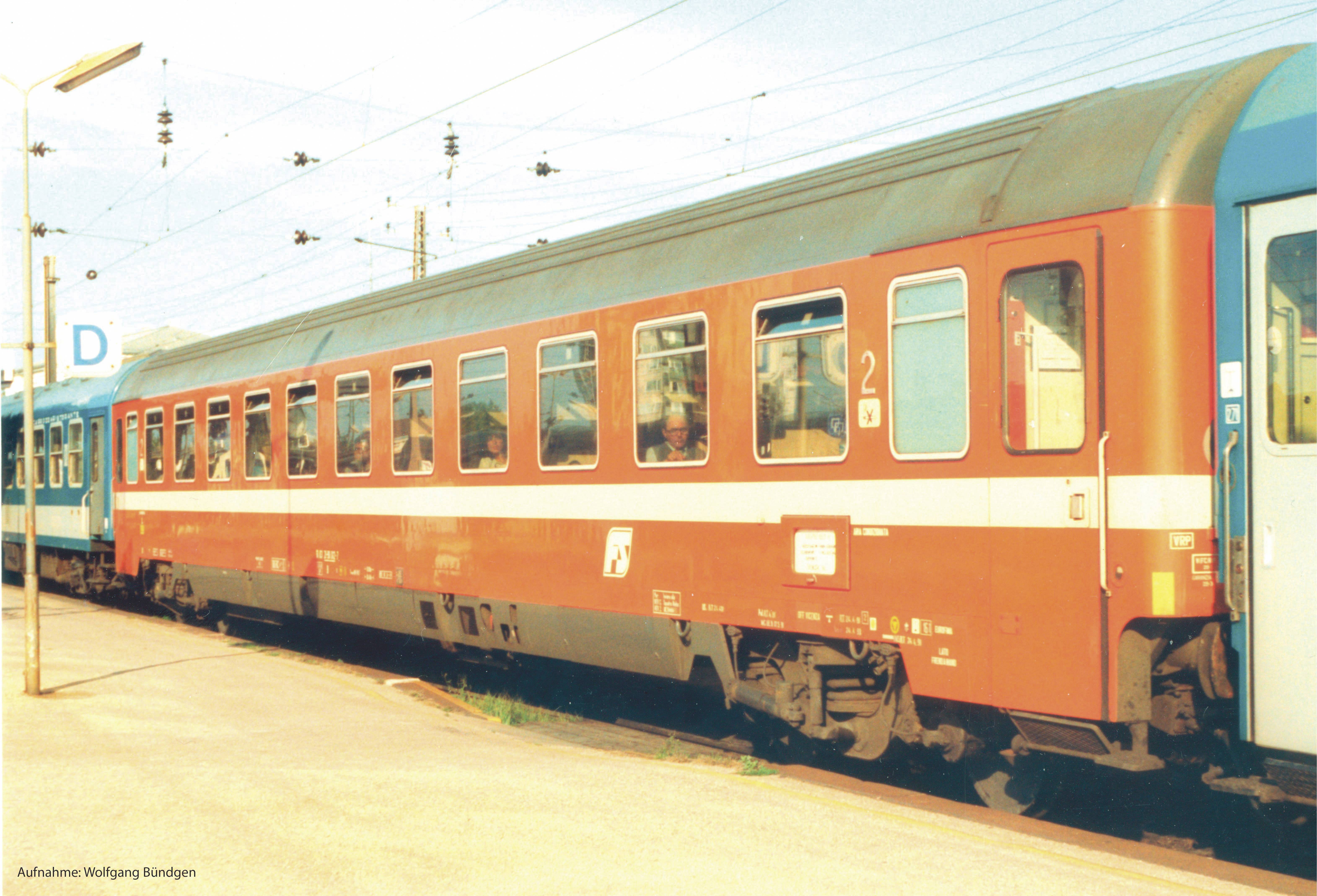 A red train carriage is standing at the railway station. People are seated inside. Overhead wires and a blue sign with 'D' are visible.
