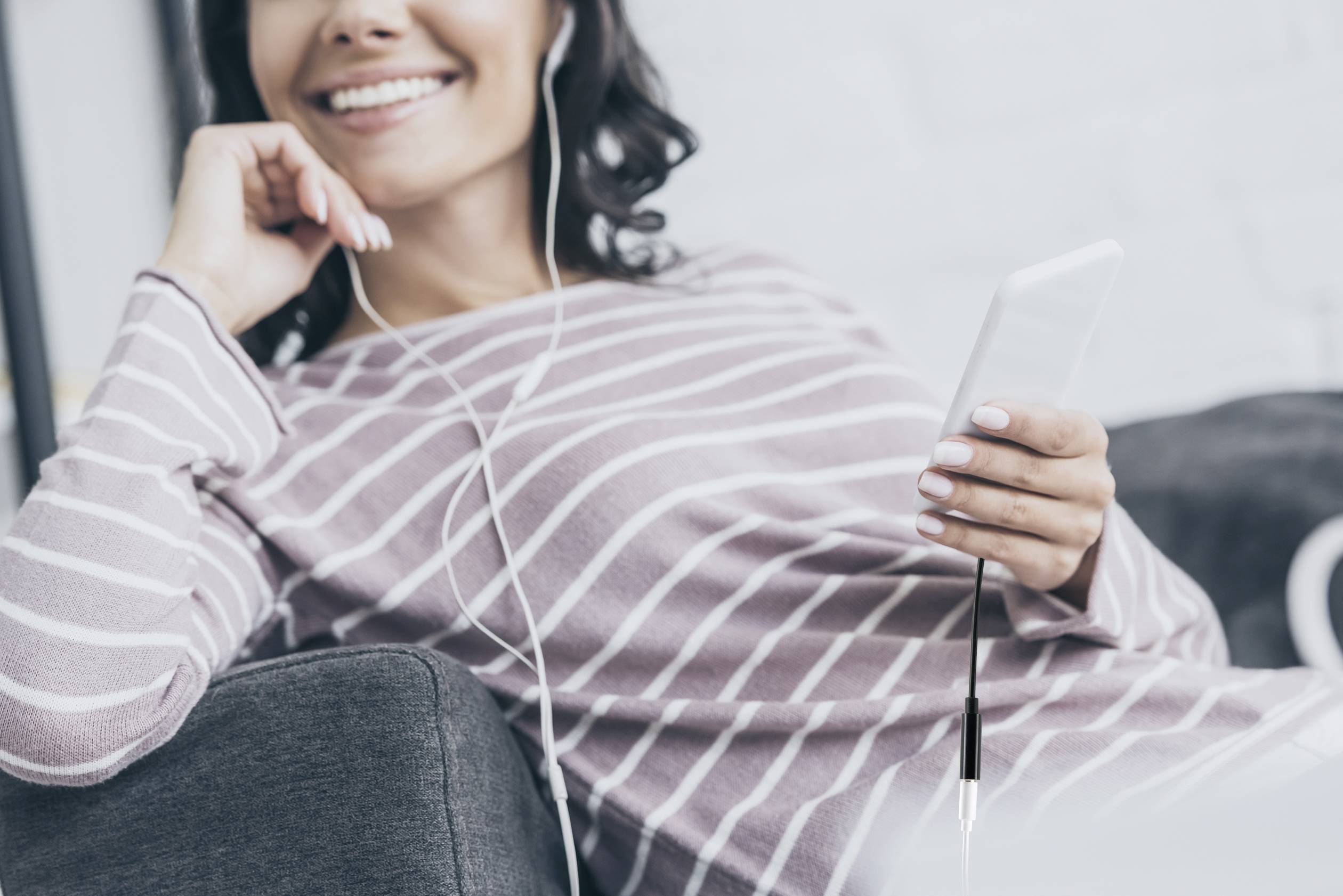 A woman sits relaxed on a sofa, smiling, listening to music through earphones and holding a smartphone in her hand.