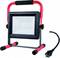 A portable LED floodlight with a red handle and black and red frame. A black cable with a plug lies beside it.