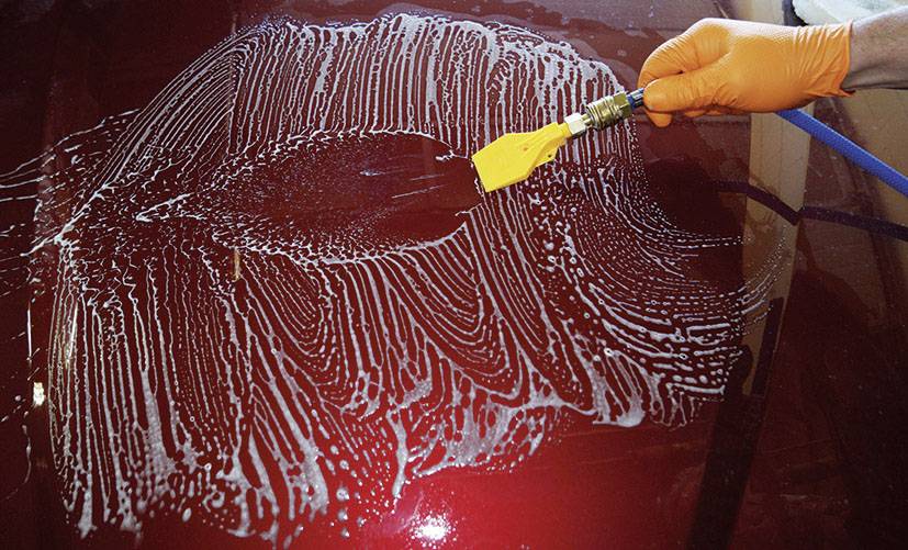A person is cleaning the bonnet of a car with a brush connected to a hosepipe. The bonnet is covered in soapsuds.