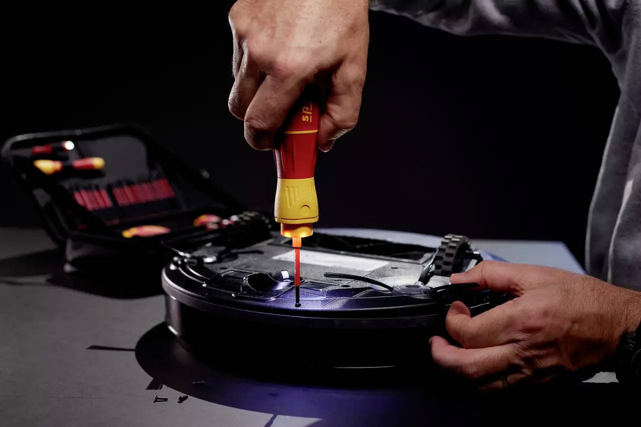 A person is repairing a robot vacuum cleaner with a screwdriver. An open toolkit lies in the background.
