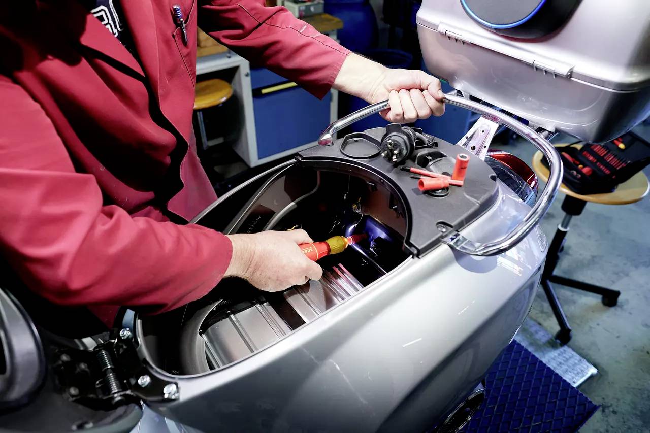 A person is working on an electric scooter. They are using tools to inspect the engine compartment at the rear of the scooter.