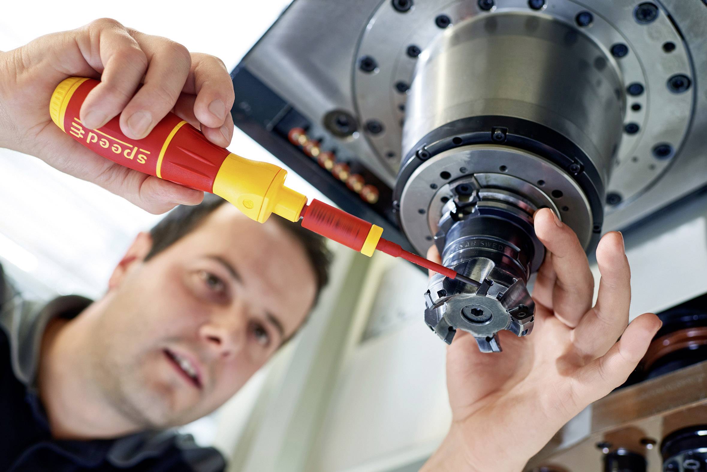 A person is adjusting a metal drill bit with a yellow screwdriver on a large machine. The focus is on precise manual work.