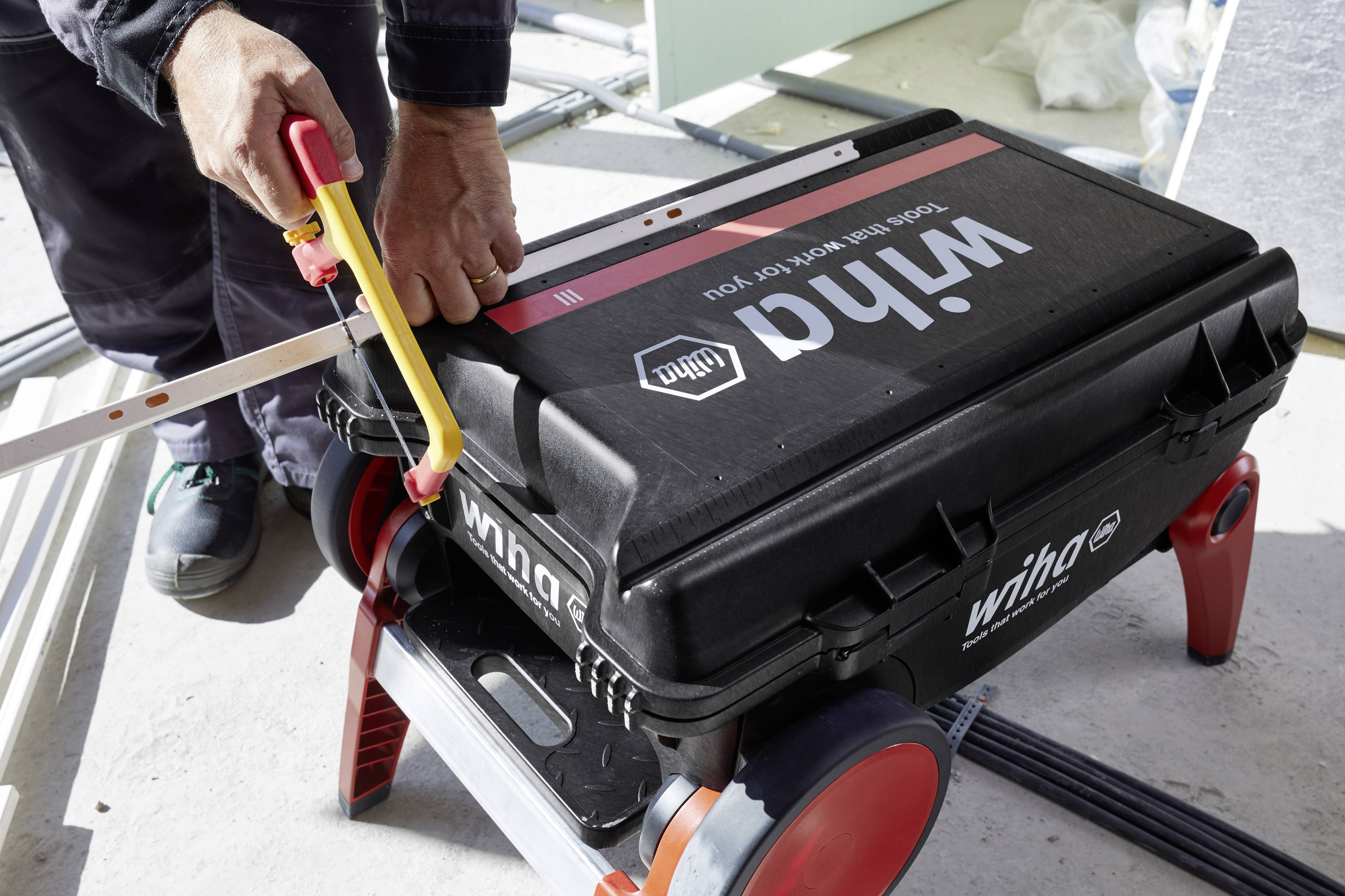 A person is using a saw to cut a piece of wood on a black Wiha toolbox with wheels.