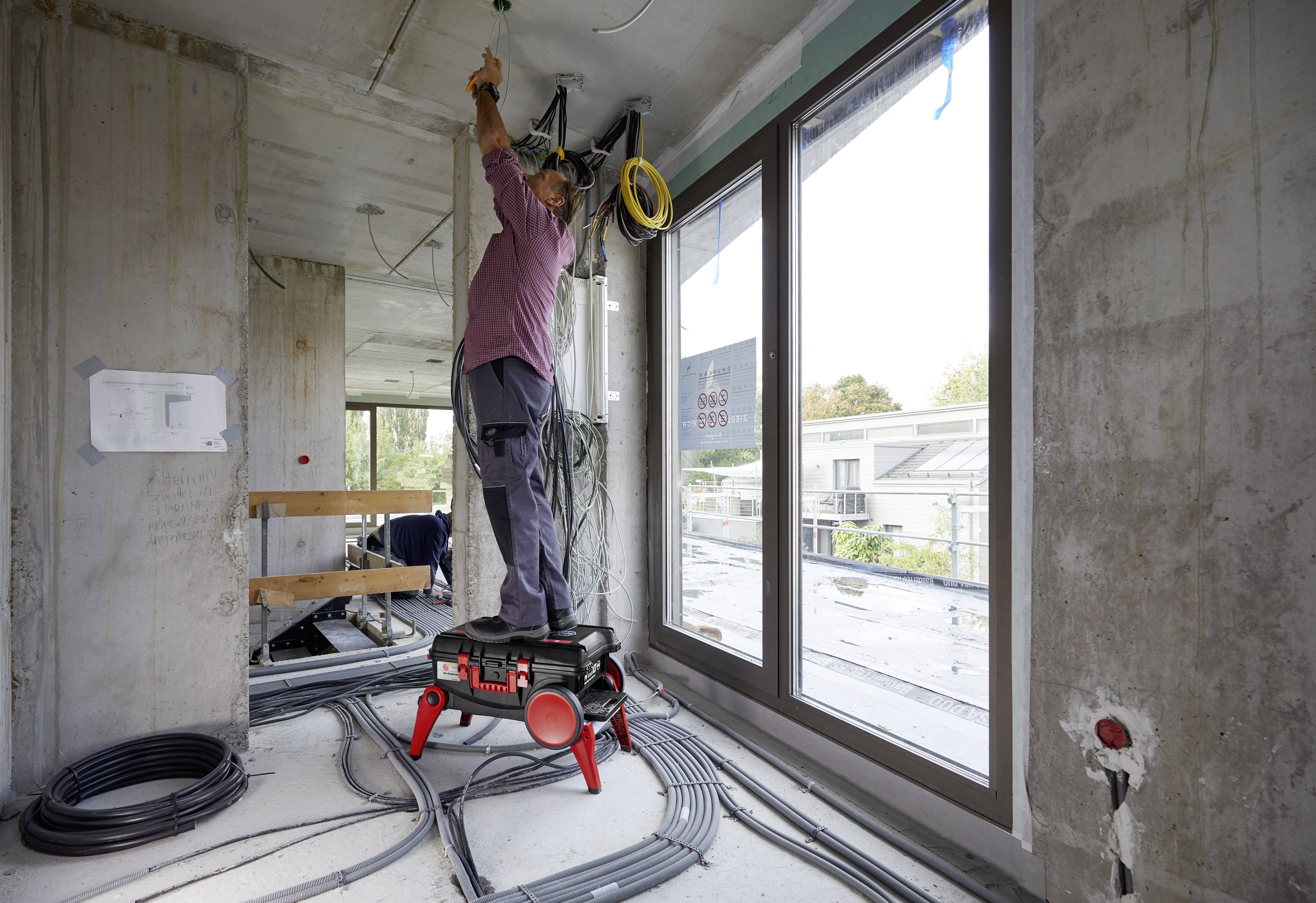 A tradesman is working on a building site, standing on a crate and installing electrical cables on the ceiling of an unfinished building.