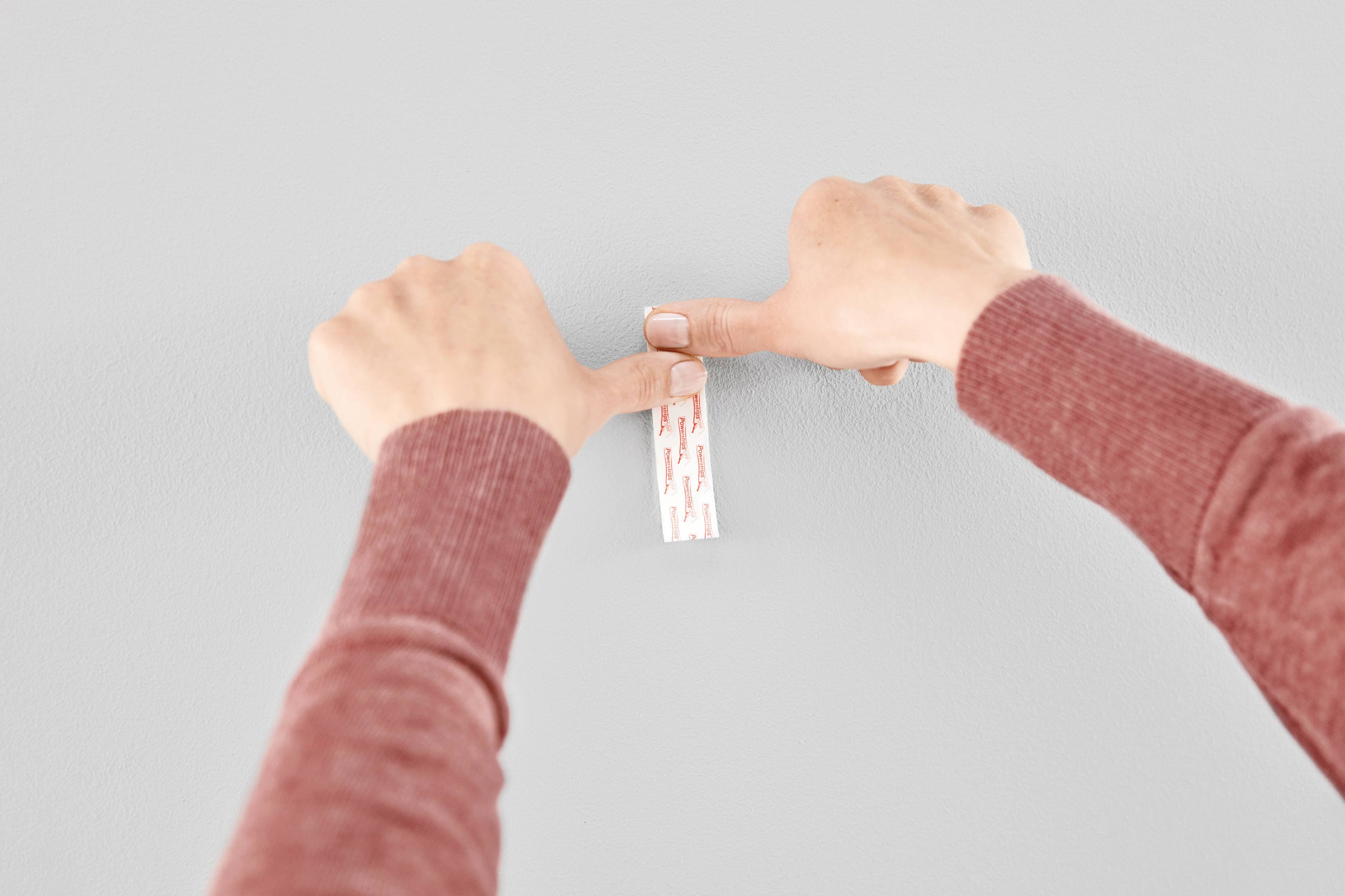 Two hands are pressing an adhesive strip onto a grey wall. A person is securing something, possibly a picture or poster.