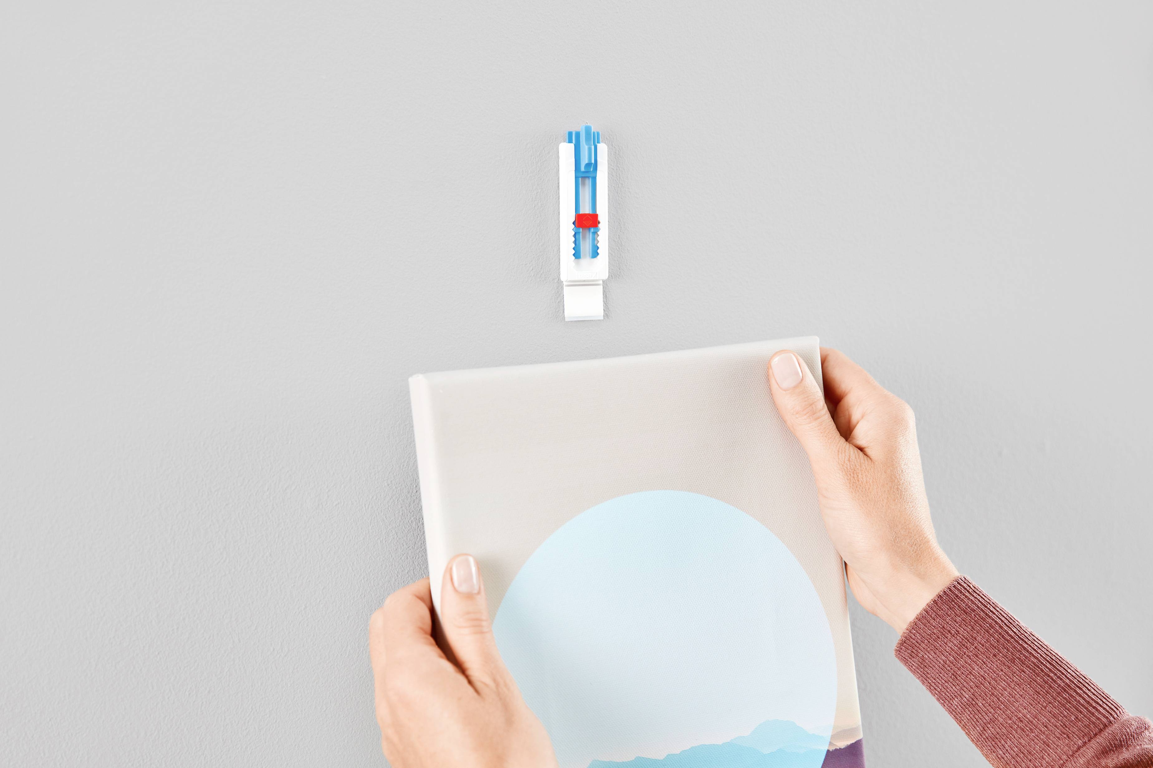 A person is hanging a picture on a grey wall using a modern hanging system that includes a blue wall plug and white hook.