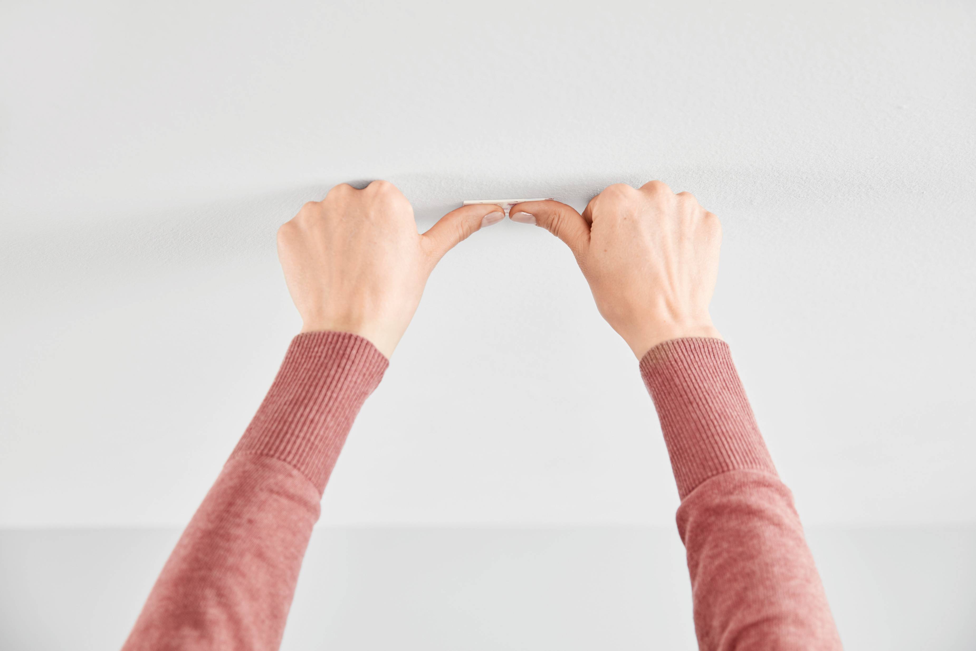 Two hands in red sleeves are securing a small object to a white ceiling, possibly an adhesive or decorative element.