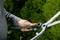 A person is holding a pair of garden secateurs in front of a background of green plants. The hand is shown close to the handle.