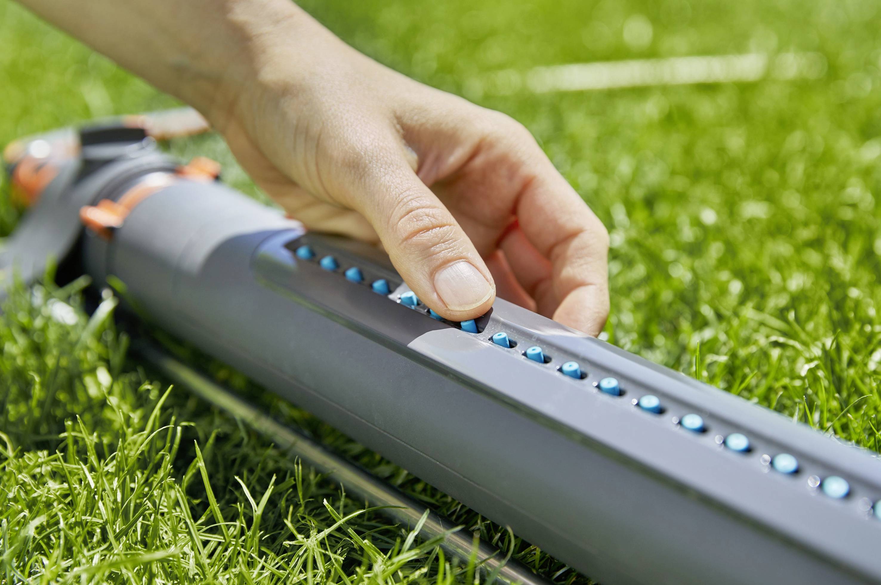 A hand adjusts a lawn sprinkler in the grass by pressing a blue button on a grey scale.
