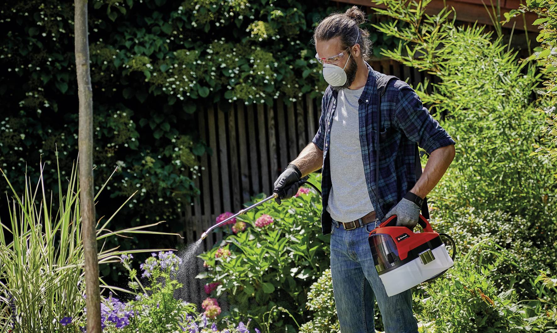 A man wearing a protective mask is spraying plants in a garden with a handheld device. Various bushes and trees can be seen in the background.