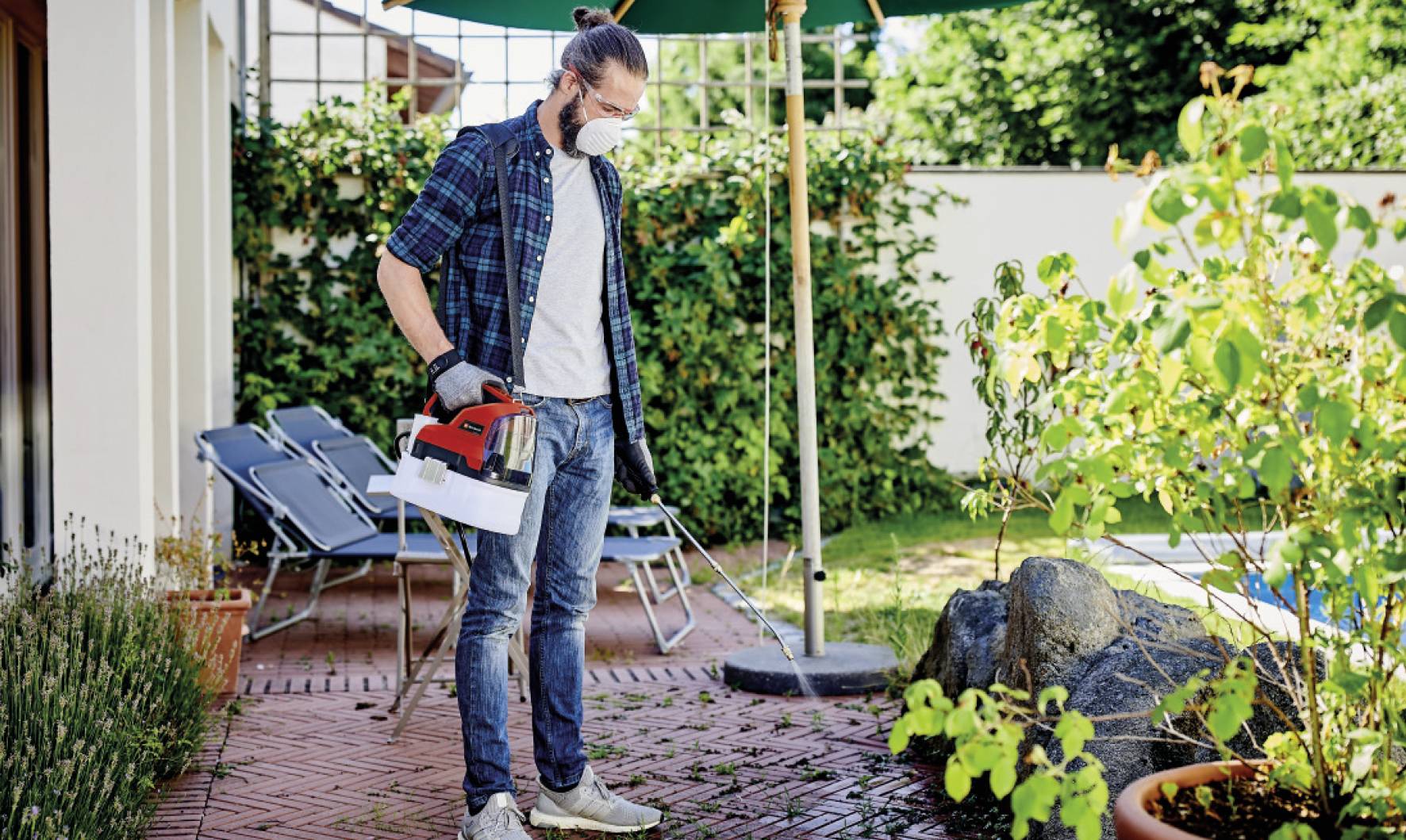 A person wearing a protective mask is watering plants in a garden with a red watering can. Garden furniture stands in the background.