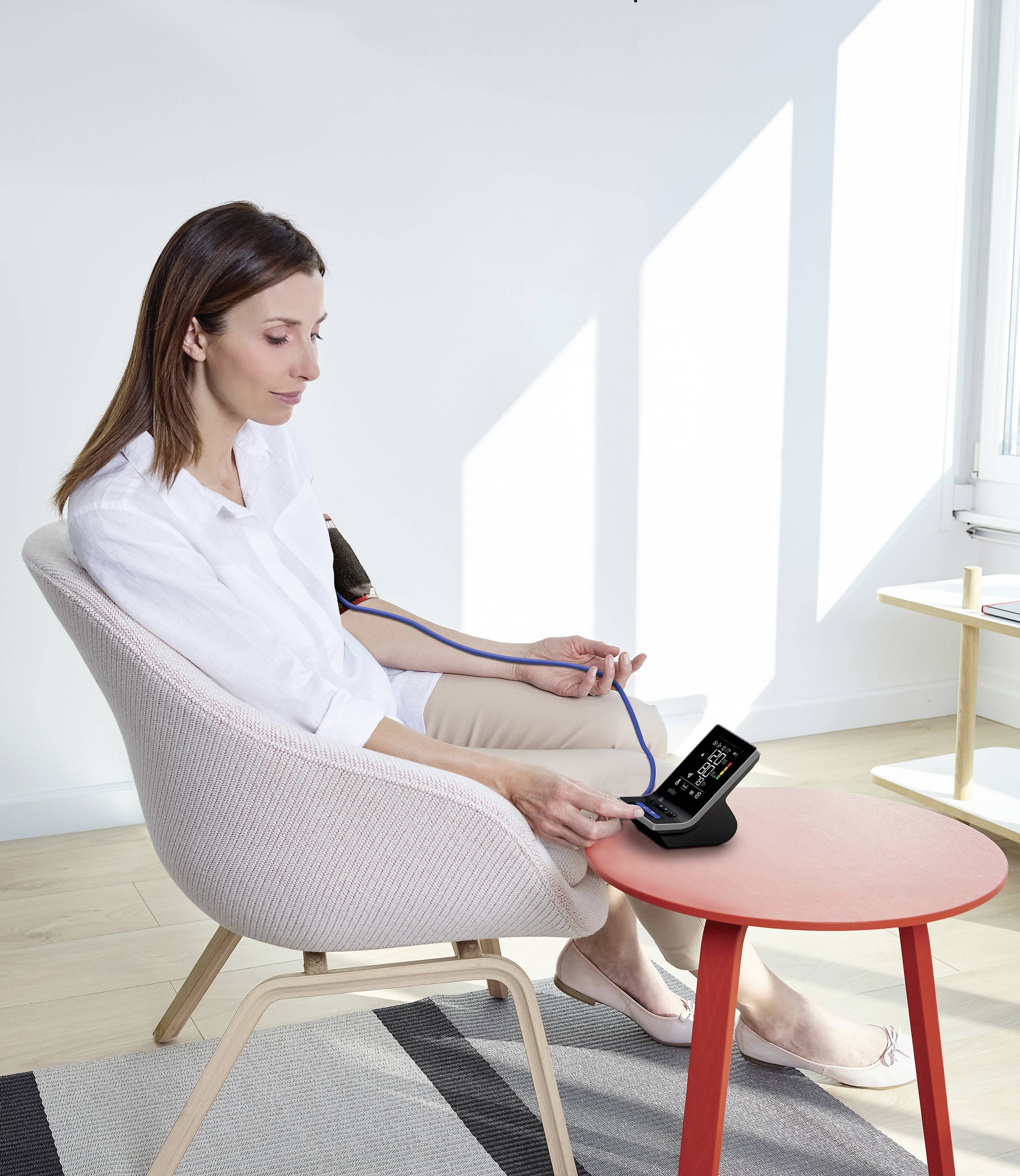 A woman is measuring her blood pressure with a digital device while sitting on a chair in a bright room.
