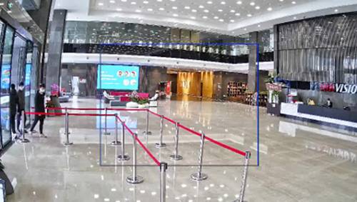 Entrance area of a modern building with a reception desk on the right. On the left, barrier tapes and people wearing masks are visible.