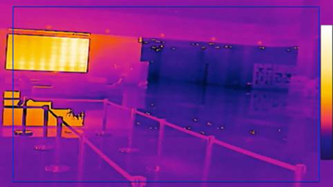 A thermal image shows an interior space with barriers in the foreground and a large bright screen on the left. The colour palette ranges from violet to yellow.