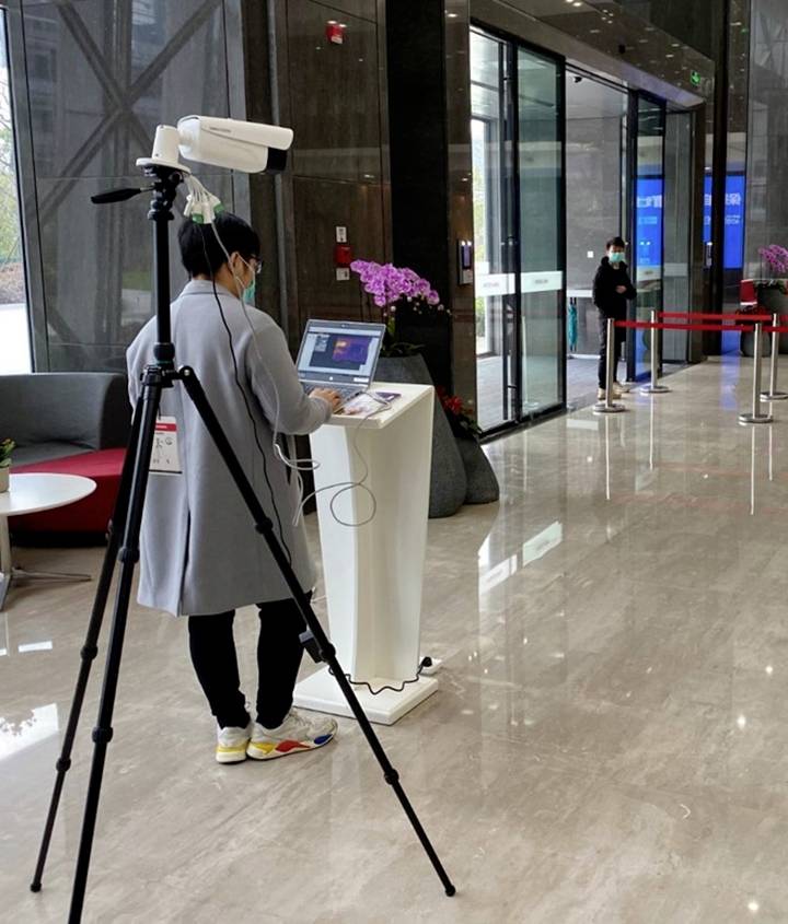 A person is standing at a lectern with a laptop, while a camera is positioned on a tripod. In the background is an entrance area.