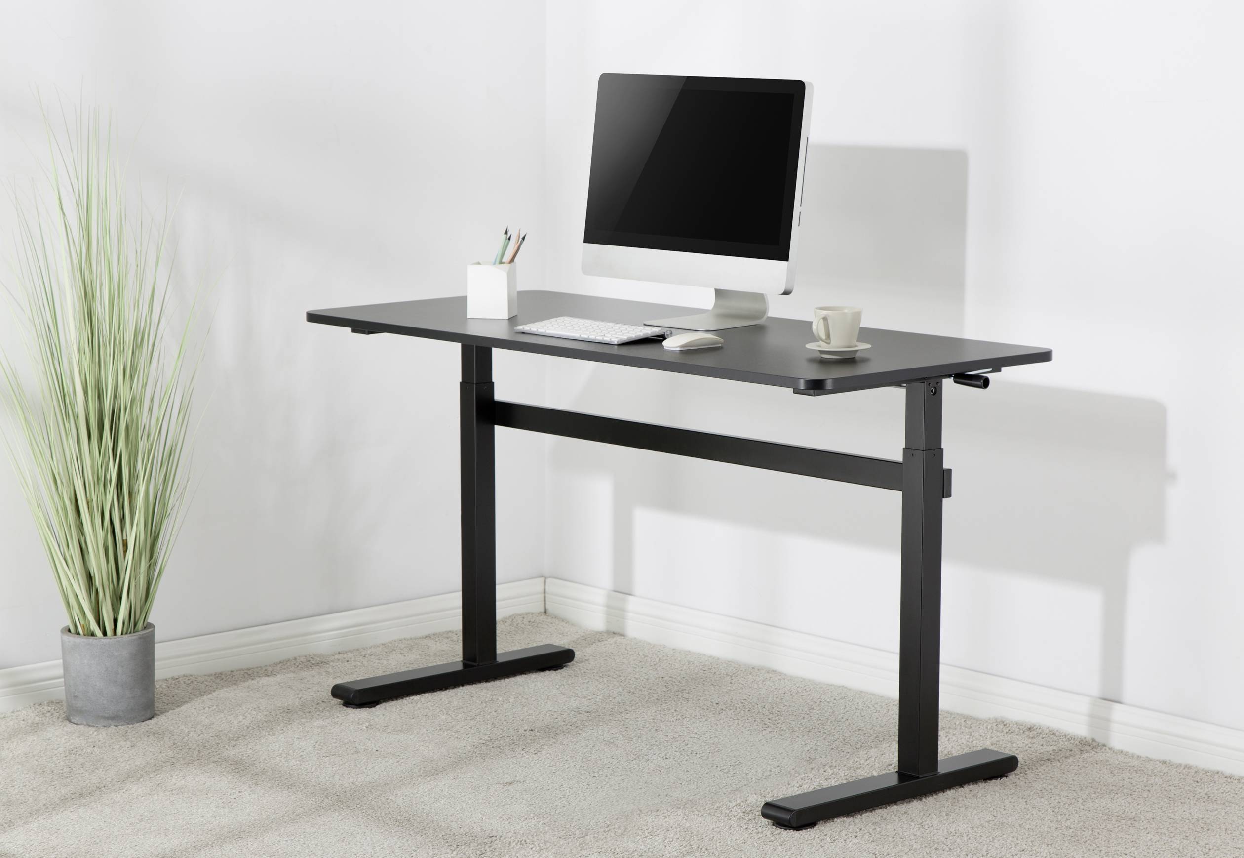 A height-adjustable desk with a black surface, featuring a monitor, keyboard, and mug. Beside it stands a plant.