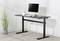 A height-adjustable desk with a black surface, featuring a monitor, keyboard, and mug. Beside it stands a plant.