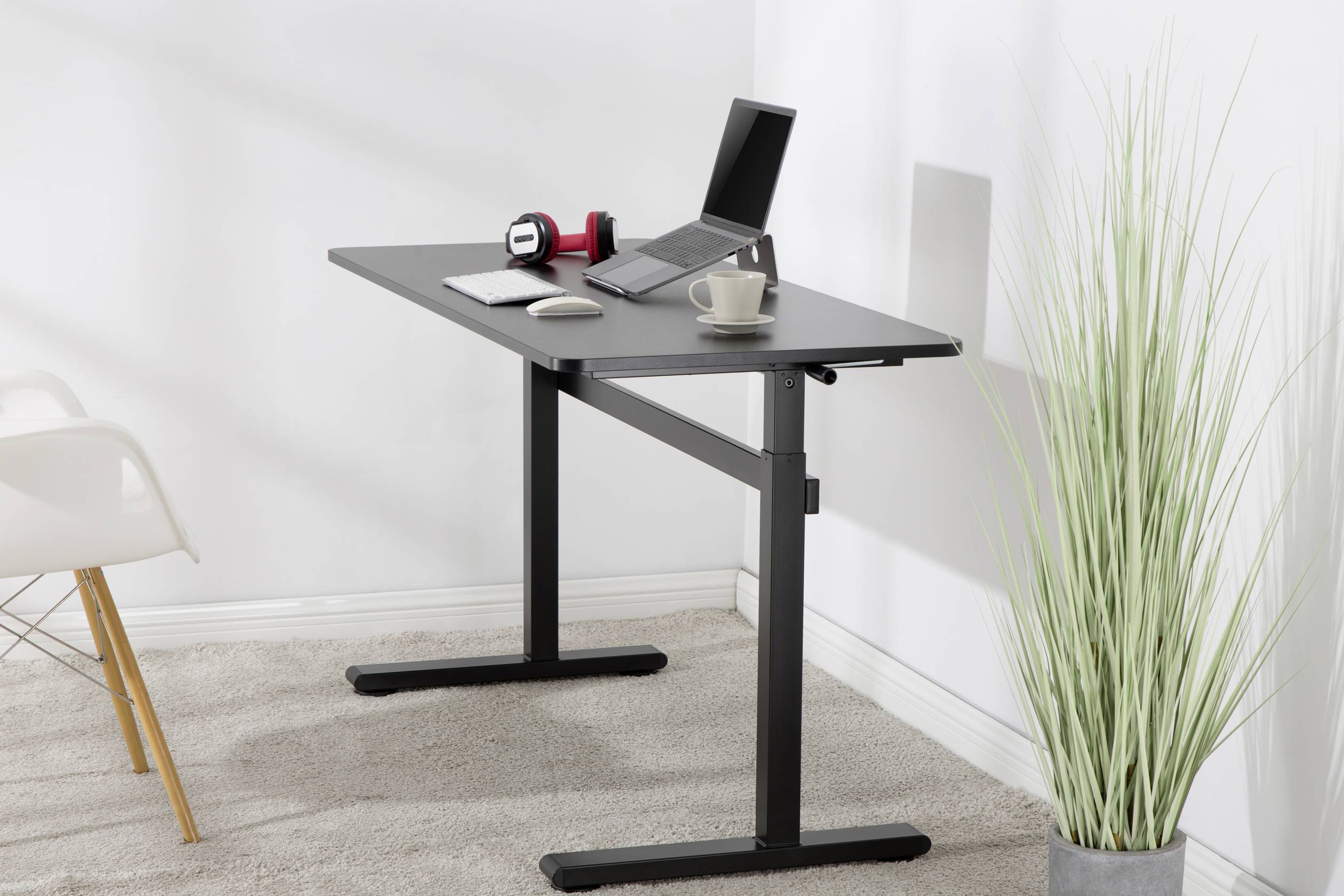 An adjustable-height, black desk with a laptop, headphones, a mug, and a mouse pad on top, sits on a beige carpet.
