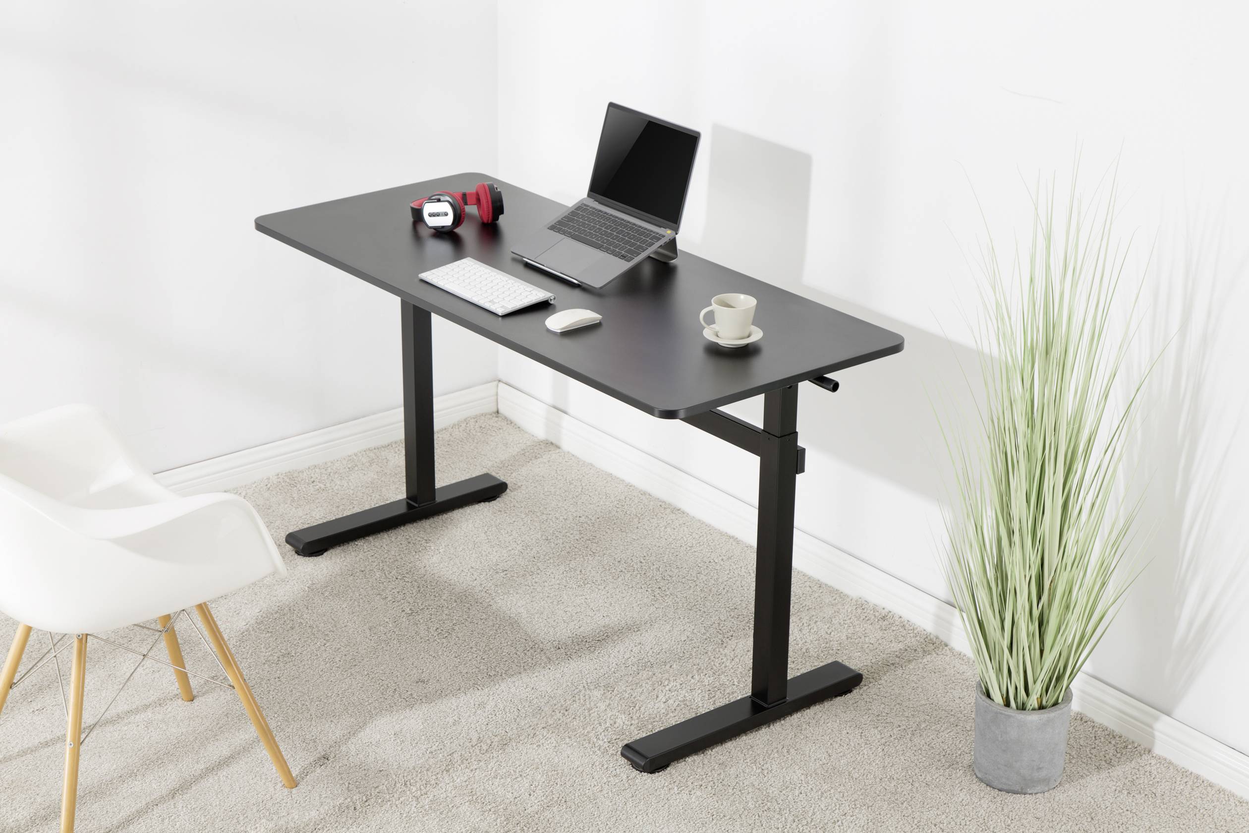 A modern desk with a laptop, keyboard, mouse, headphones and a mug. Next to it stands an office chair and a plant on carpeted flooring.