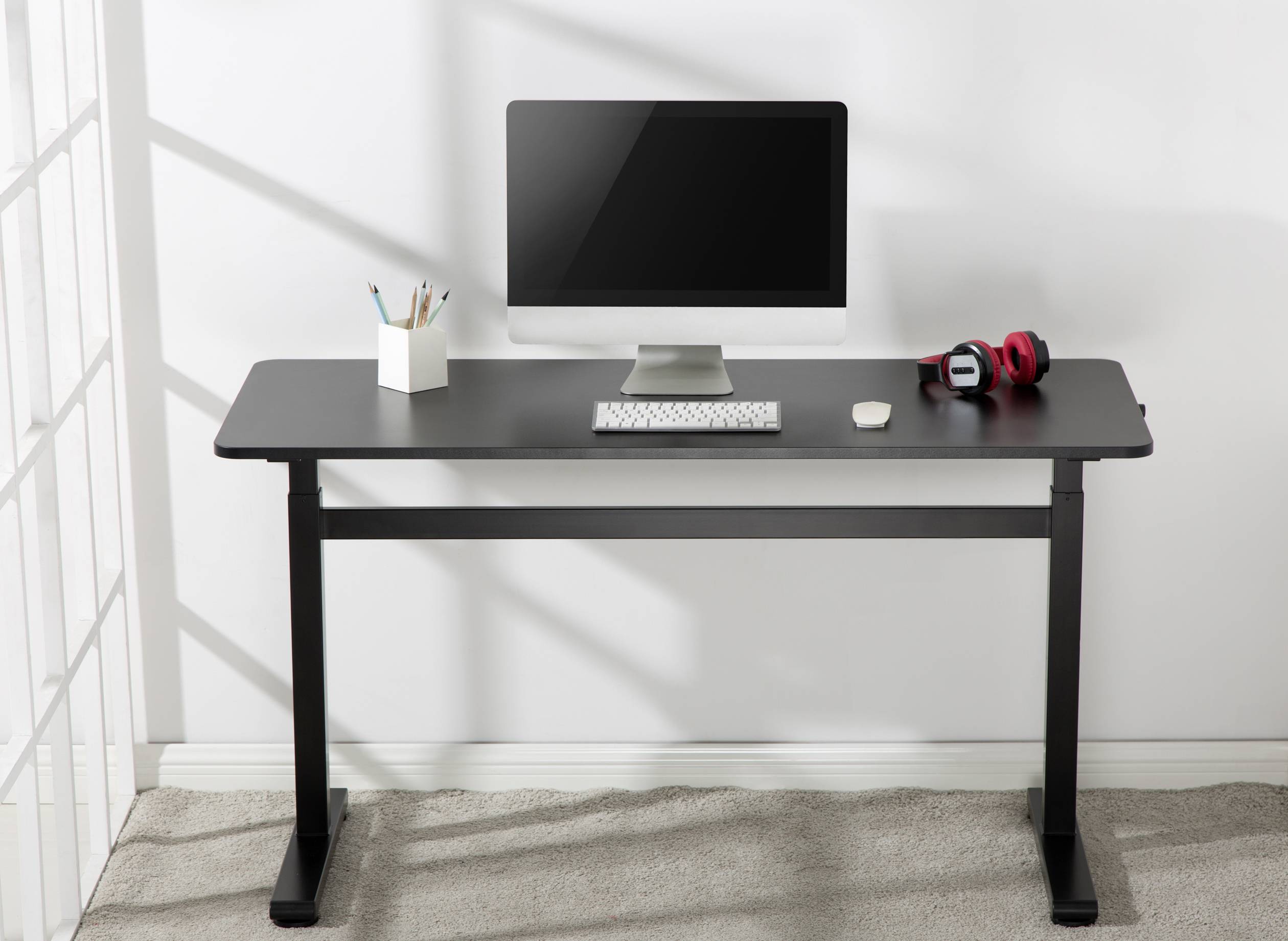 A black desk with a computer, keyboard and mouse. Next to it stands a mug with pens and red headphones. Background: white room.