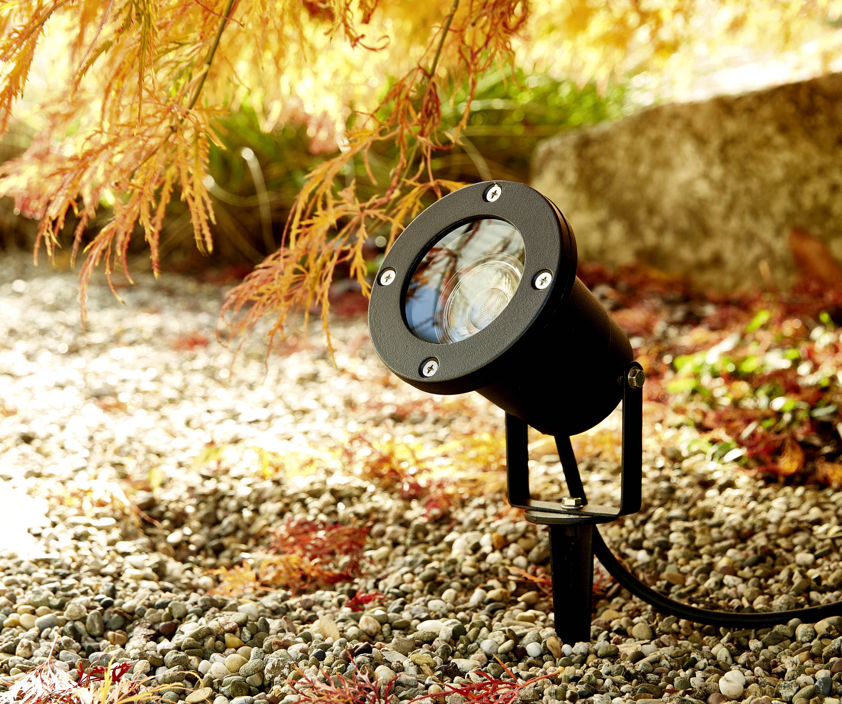 A black outdoor spotlight on a gravel-covered ground, surrounded by orange autumn leaves beneath a tree.