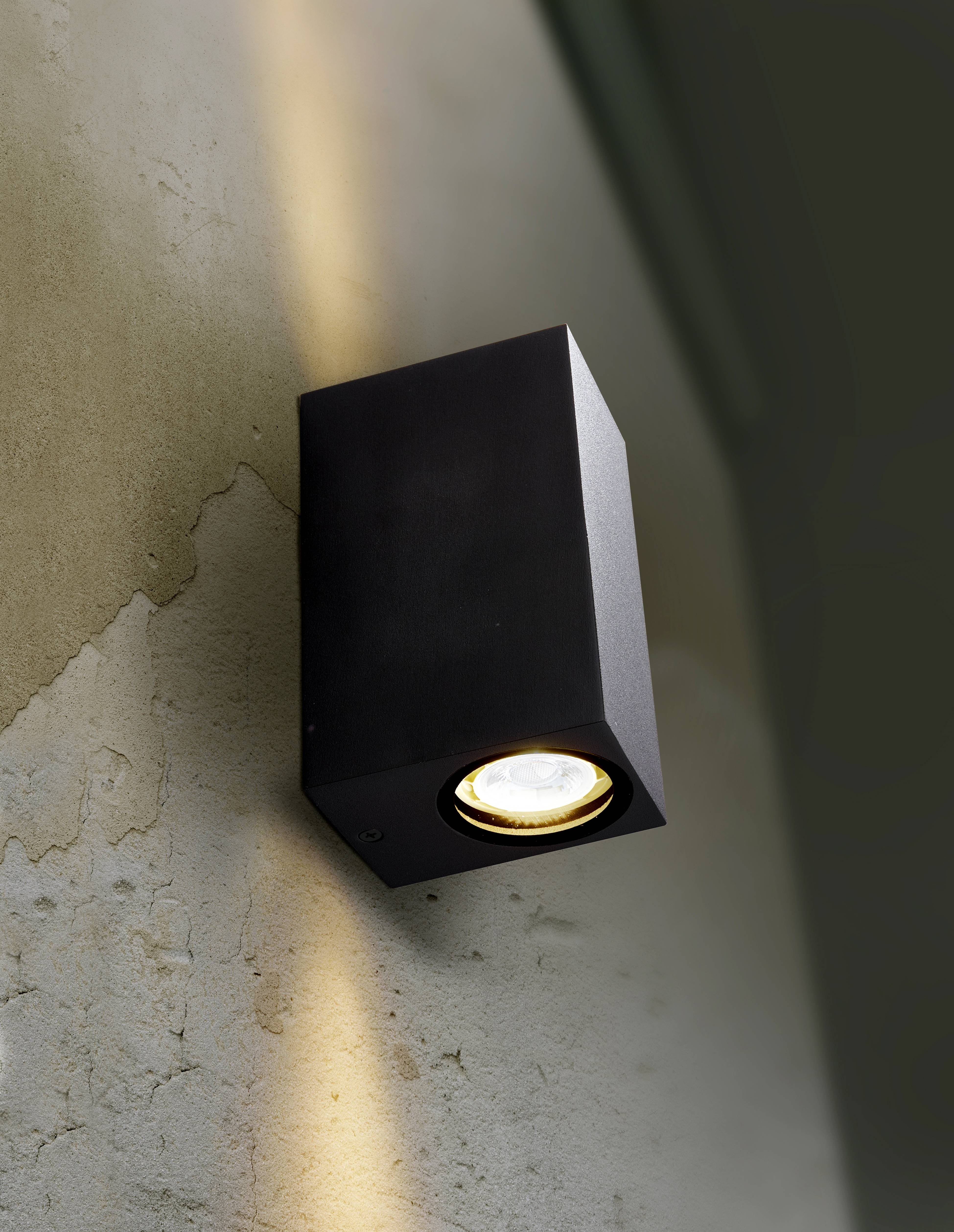 Wall light with a modern, black casing illuminates a rough, beige wall with a warm downward beam of light.
