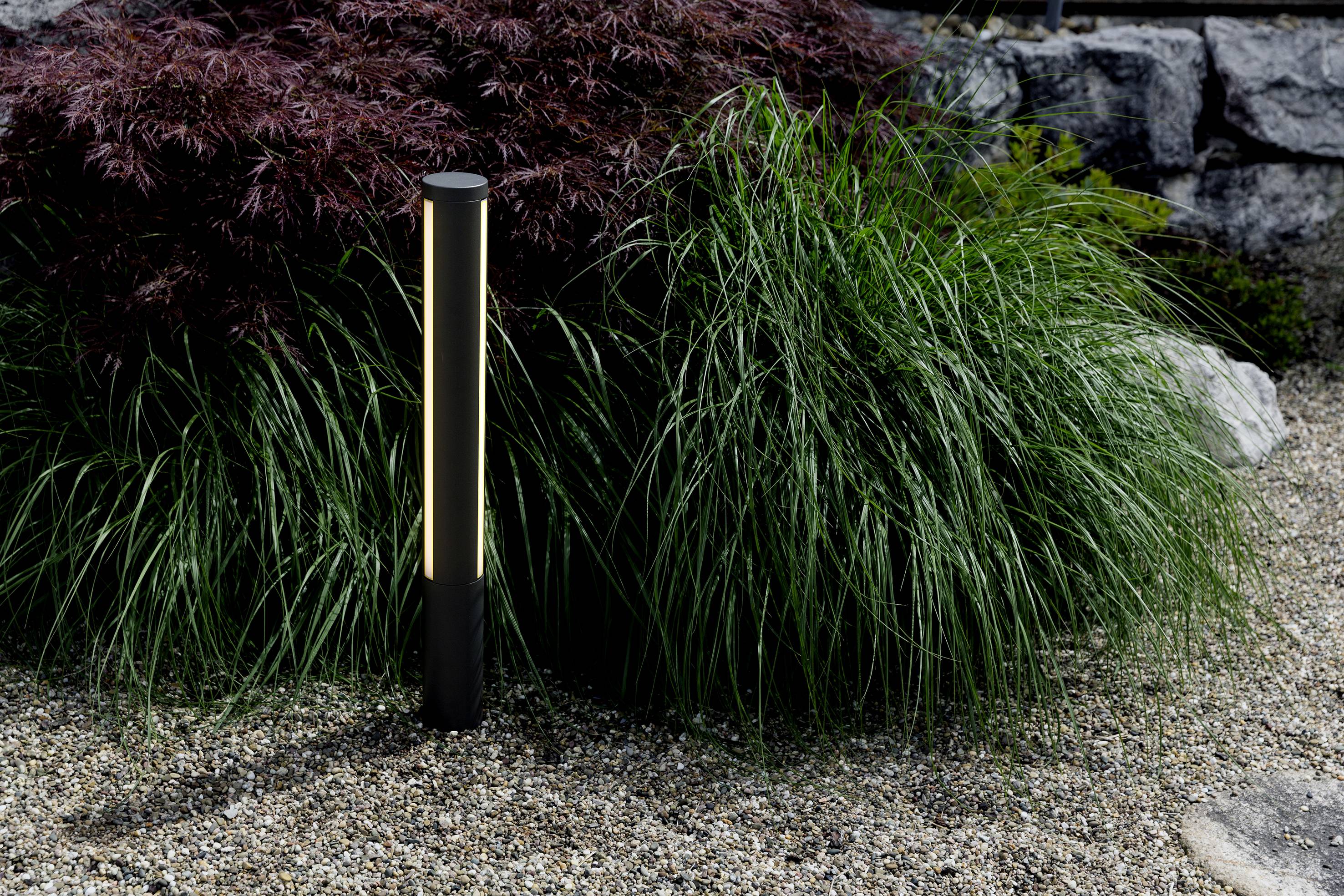 A slender, illuminated outdoor light stands on a gravel bed beside green ornamental plants and a stone wall in the background.
