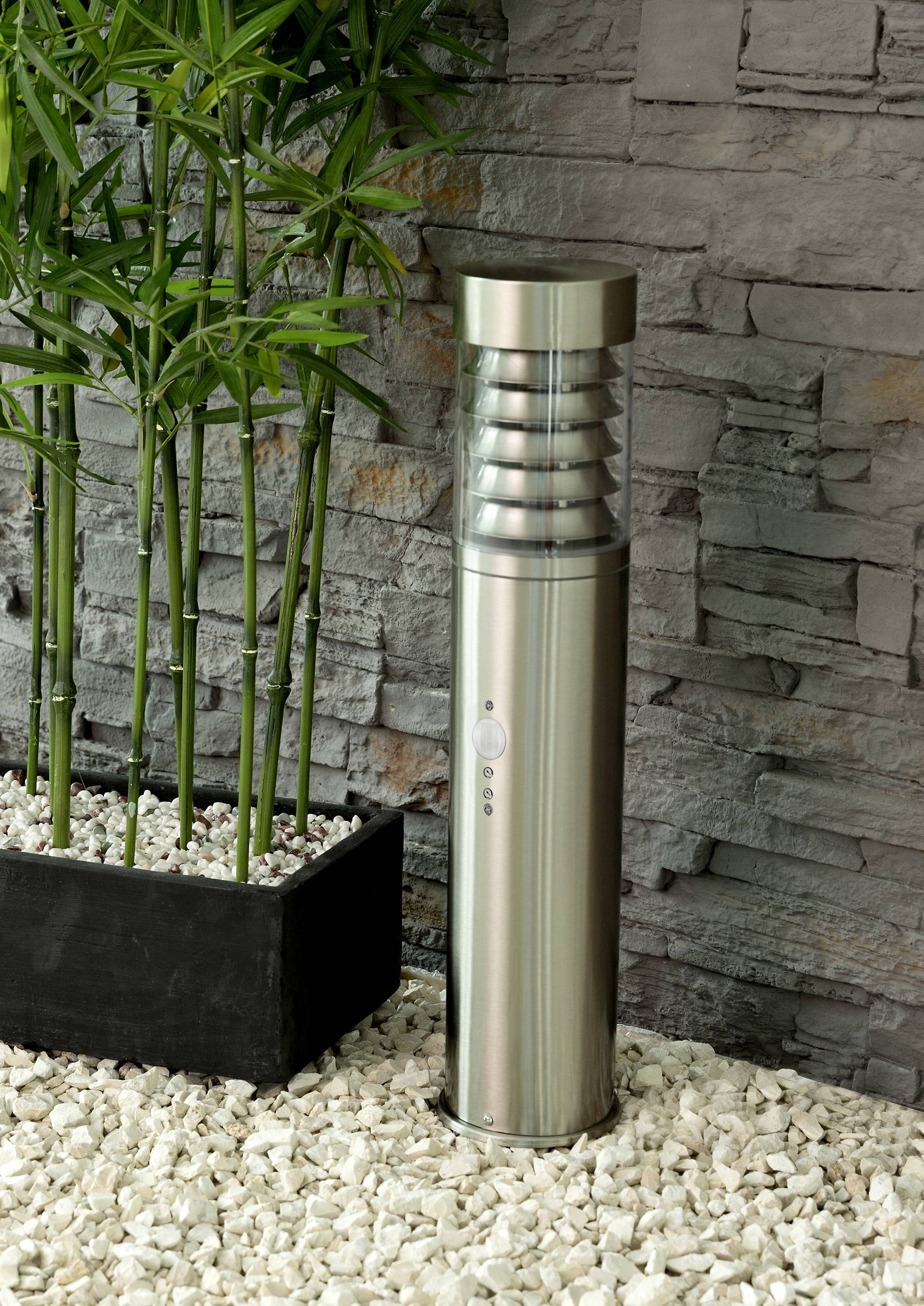 Silver outdoor light with modern design stands on white gravel beside green bamboo in a planter in front of a grey stone wall.