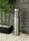 Silver outdoor light with modern design stands on white gravel beside green bamboo in a planter in front of a grey stone wall.