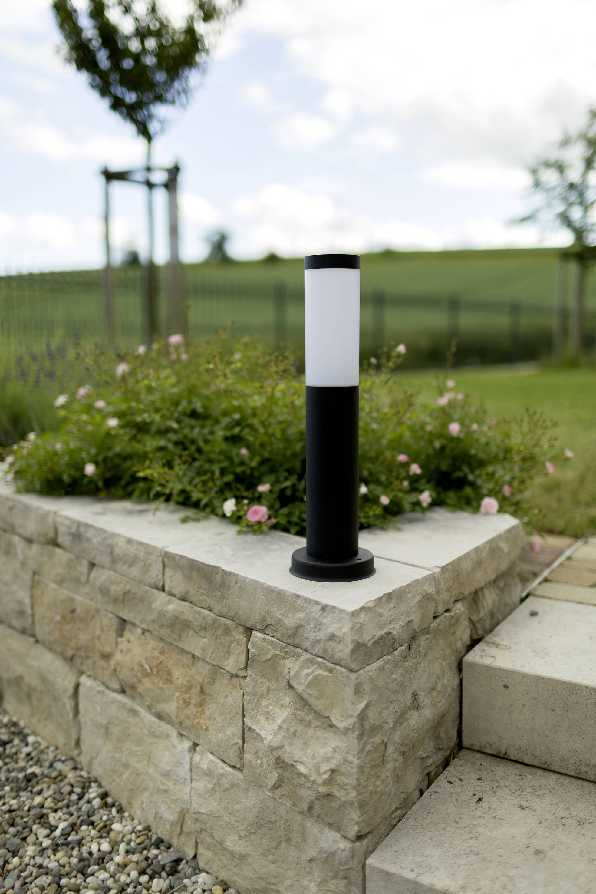 A modern, black garden light stands on a stone wall, surrounded by green plants and a field in the background.