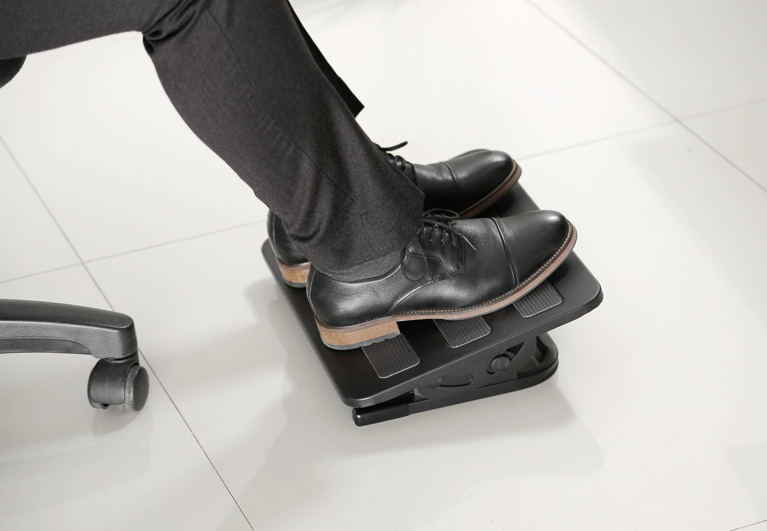 A person is sitting on an office chair and using a black footrest to elevate their feet. The support has an adjustable design.