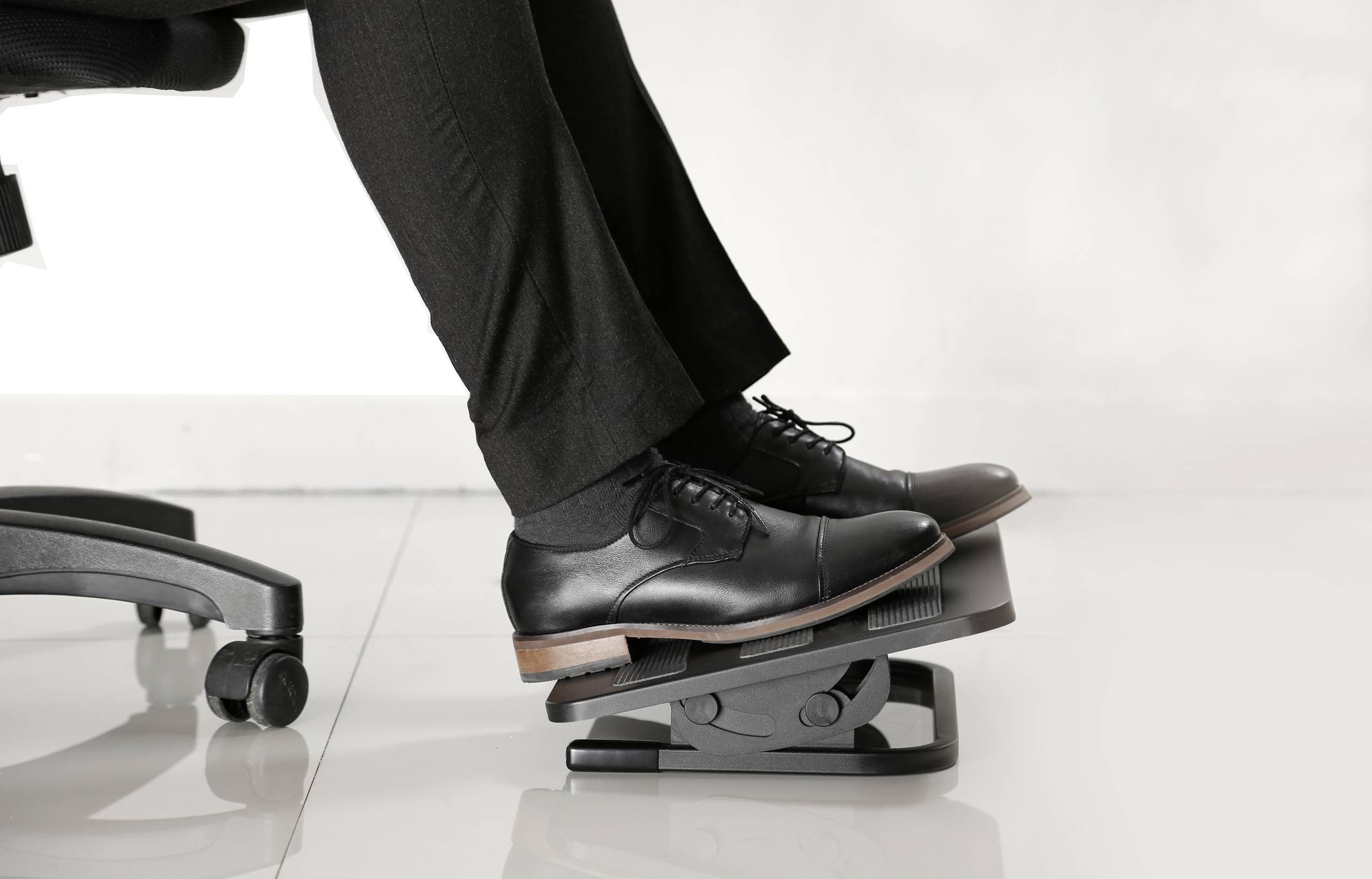 A person wearing smart shoes is using a footrest under a desk chair to ensure ergonomic comfort while sitting.