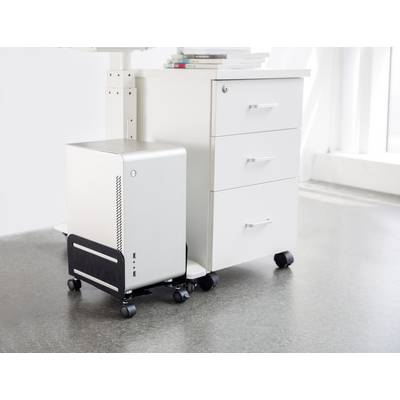 A silver computer on a stand sits beside a white mobile pedestal with three drawers on a concrete floor in a bright room.