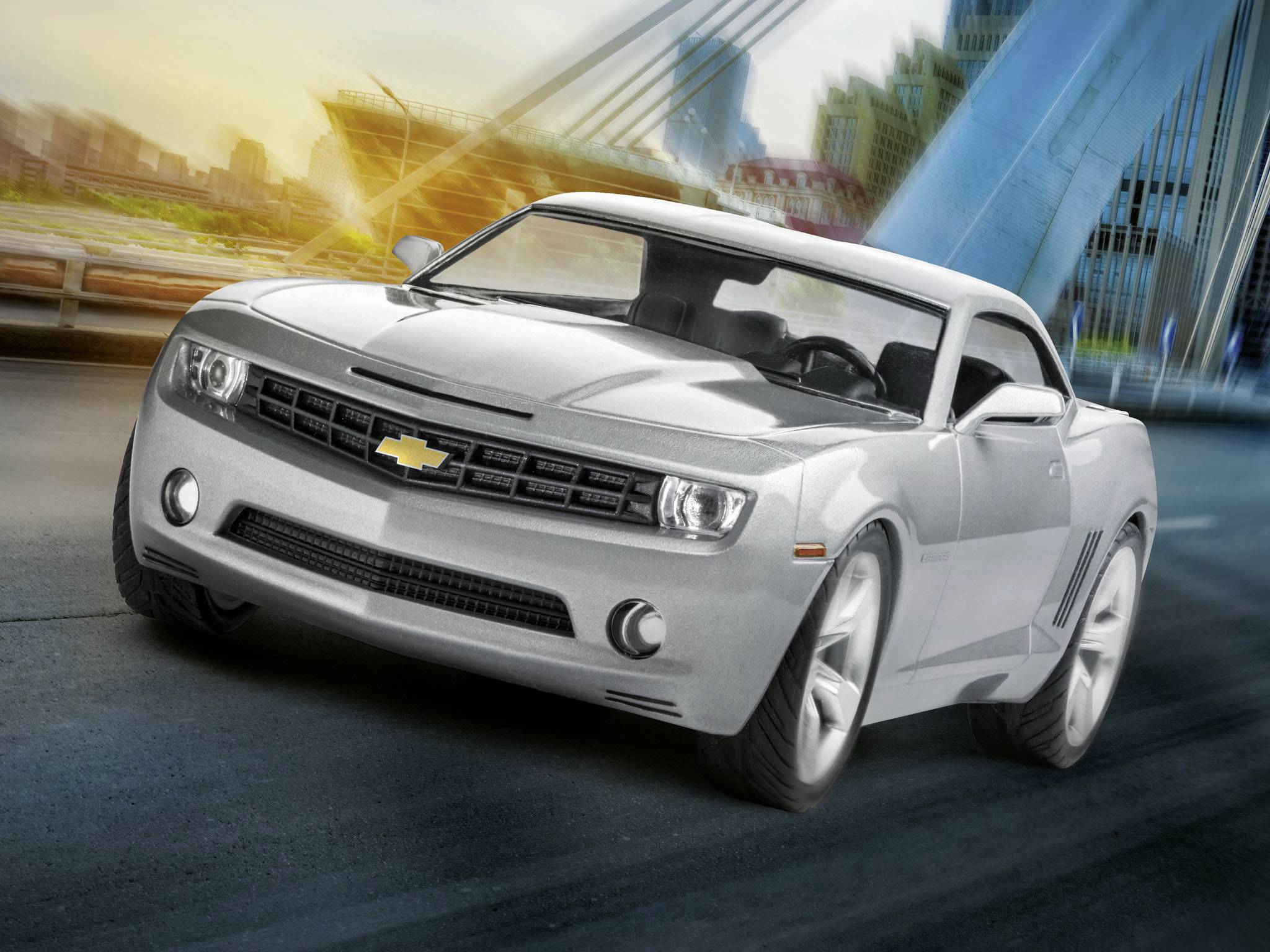 A silver-coloured car drives on a bridge in an urban setting at sunset. Modern buildings are visible in the background.