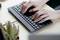 Hands typing on a black computer keyboard on a desk, with a small cactus in a pot beside it.