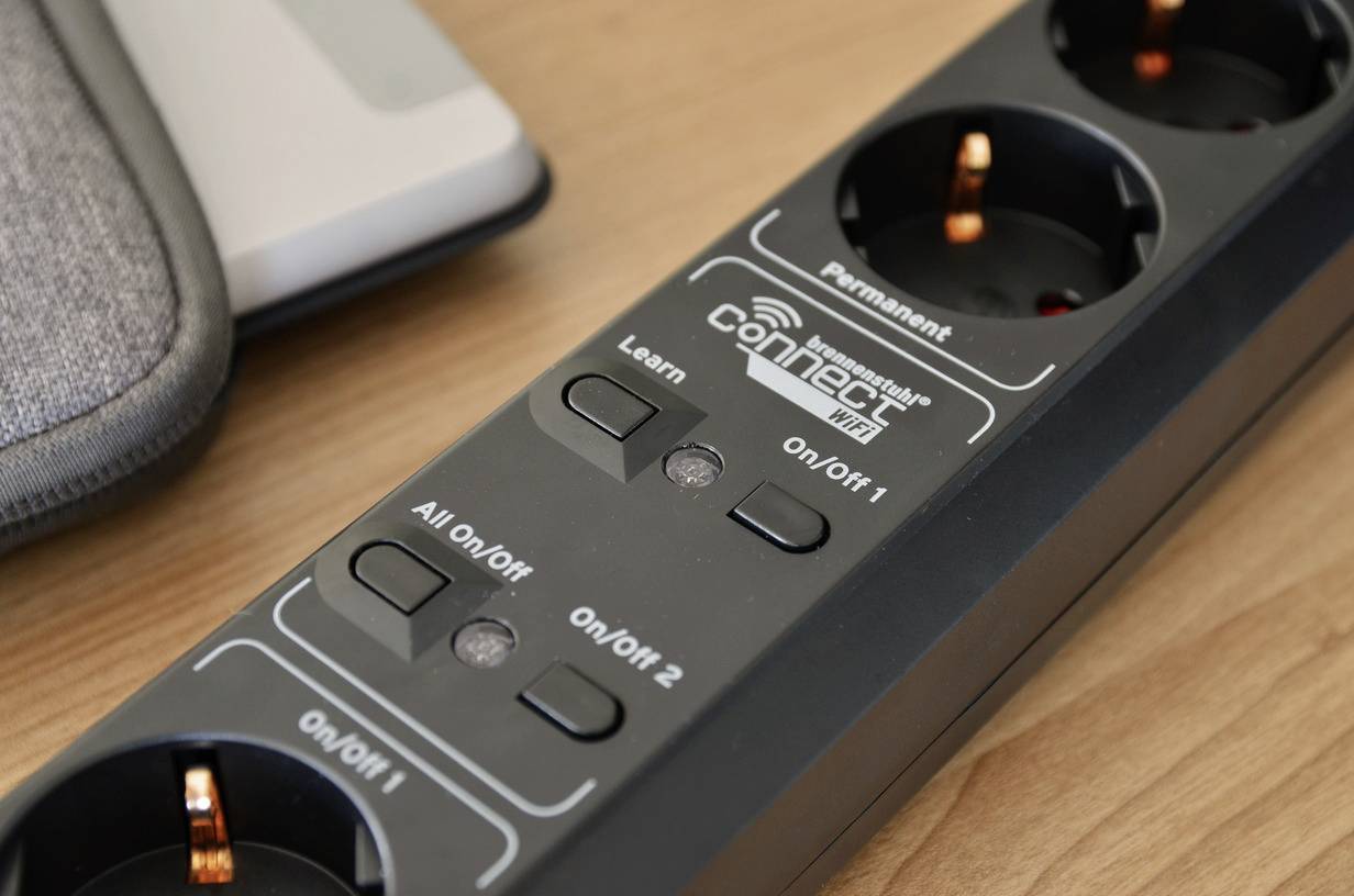 Multi-socket extension with Wi-Fi control buttons 'On/Off 1', 'On/Off 2', 'Learn', and 'Permanent' on a wooden table.