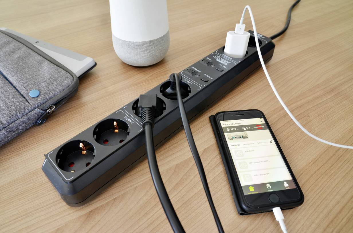A black multi-socket extension lead with several devices plugged in, including a smartphone being charged via a USB cable.