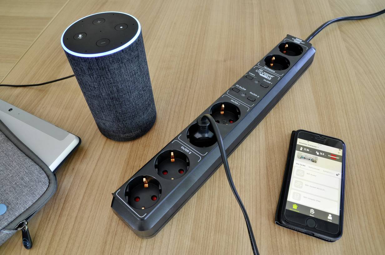 A voice assistant, a power strip and a smartphone with a smart home app lie on a table, showing interconnected technology.