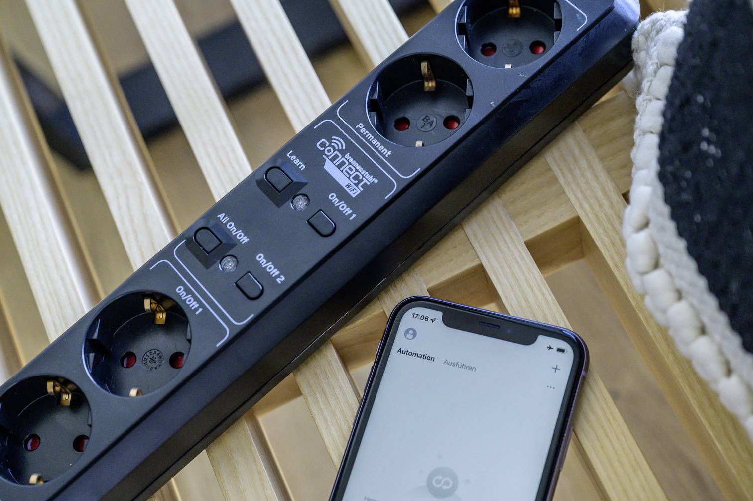 A smartphone displays a home automation app. Next to it, a multi-socket extension lead rests on a wooden chair.