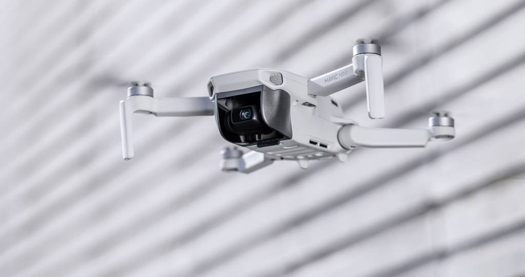 A white drone hovers in mid-air against a striped background, poised for recording or surveillance.