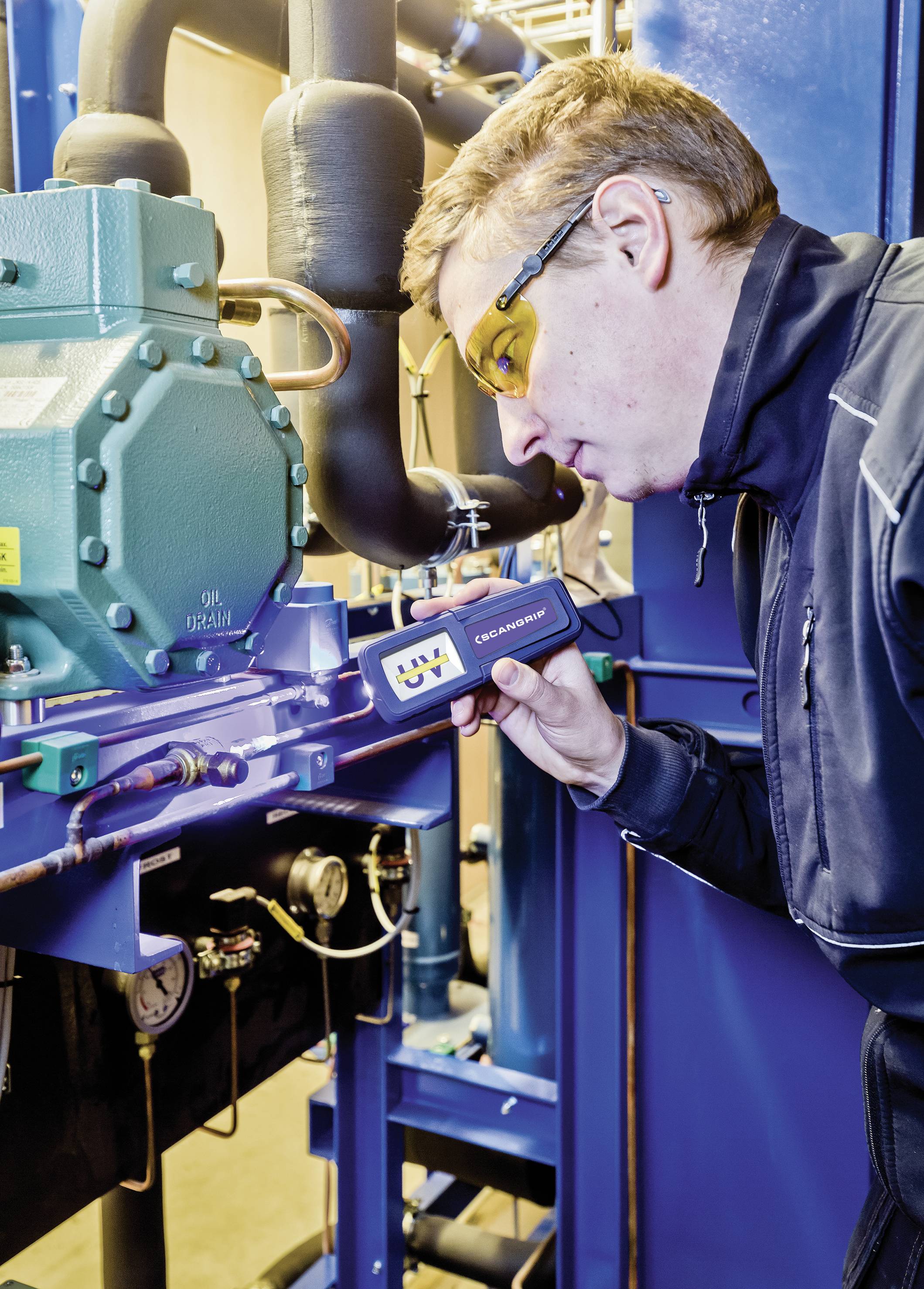 A man is using a UV leak detection device on an industrial machine, wearing safety glasses and a jacket in a workshop environment.