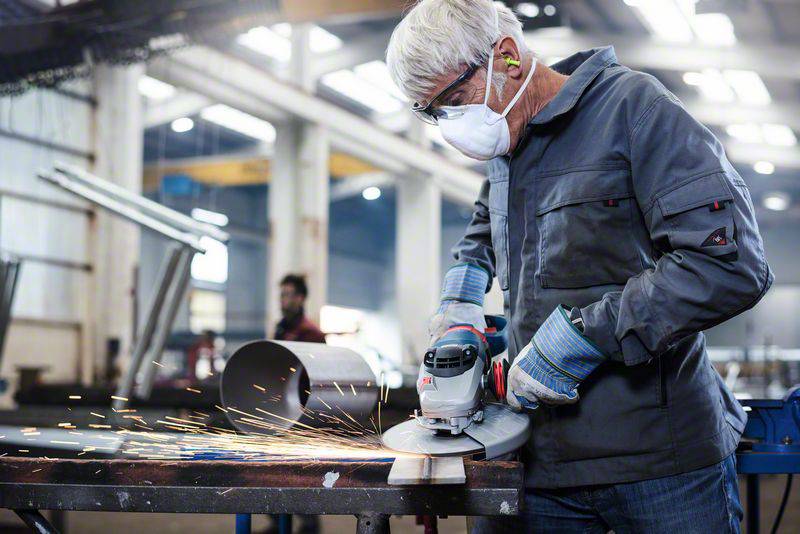An older man wearing a protective mask and glasses grinds metal in a workshop. Sparks fly as he grinds the surface.