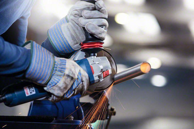 A worker in protective clothing grinds a metal pipe with an angle grinder, sparks flying.