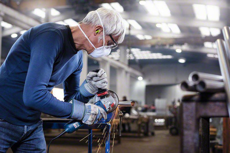 An older man wearing safety glasses, a mask, and earplugs is grinding metal in a workshop. Sparks are flying from the grinding tool.