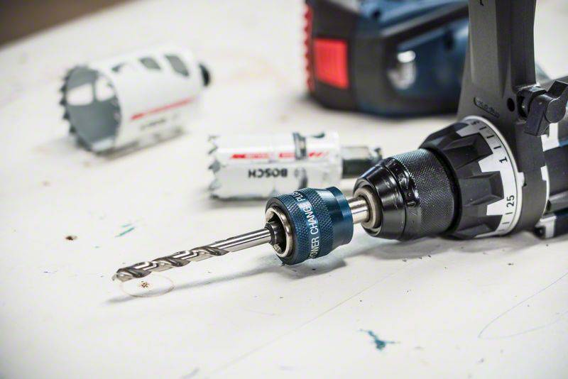 Drill with a hole saw attachment inserted, surrounded by additional drilling accessories on a workbench.