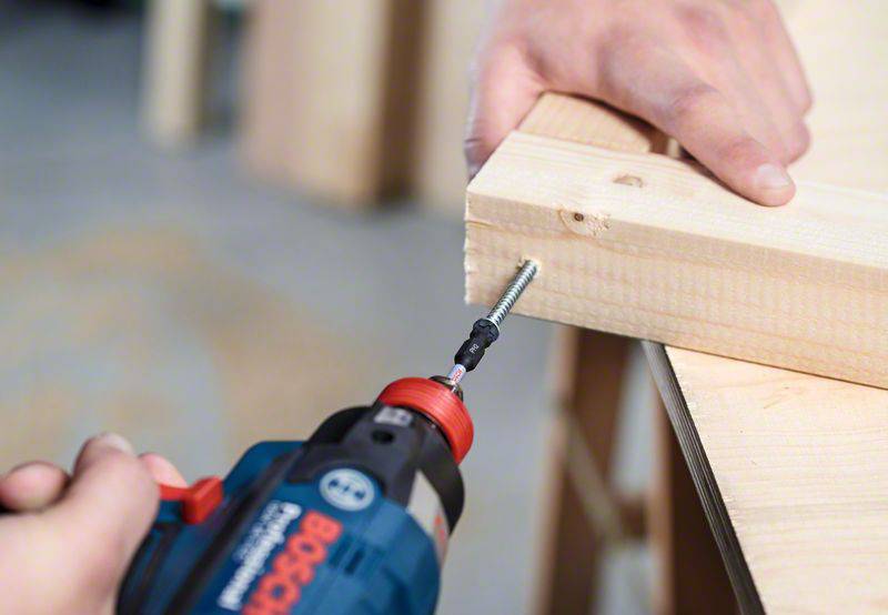 A person is drilling a screw into a wooden board on a workbench. A hand is holding the board steady in place.